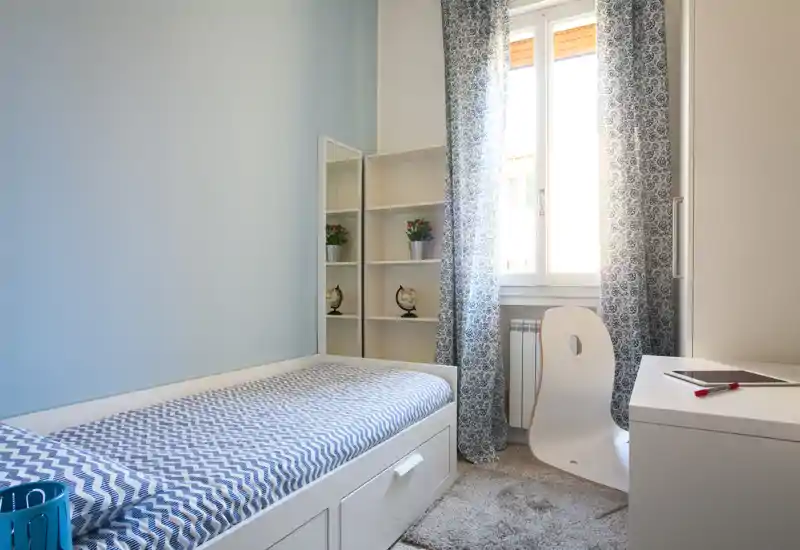 Camera singola luminosa con letto, scrivania e scaffalatura — luce naturale dalla finestra e arredi blu curati, perfetta per uno studente o un ospite.