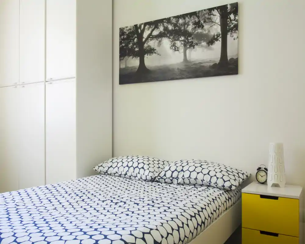 Dormitorio moderno y luminoso con cama doble con ropa de cama estampada, mesita de noche amarilla y cuadro minimalista: limpio y luminoso.