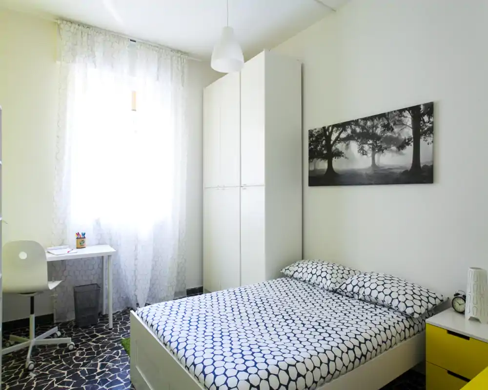 Dormitorio moderno y luminoso con cama doble, ropa de cama estampada, armario y un pequeño escritorio junto a la ventana — limpio, minimalista y acogedor.