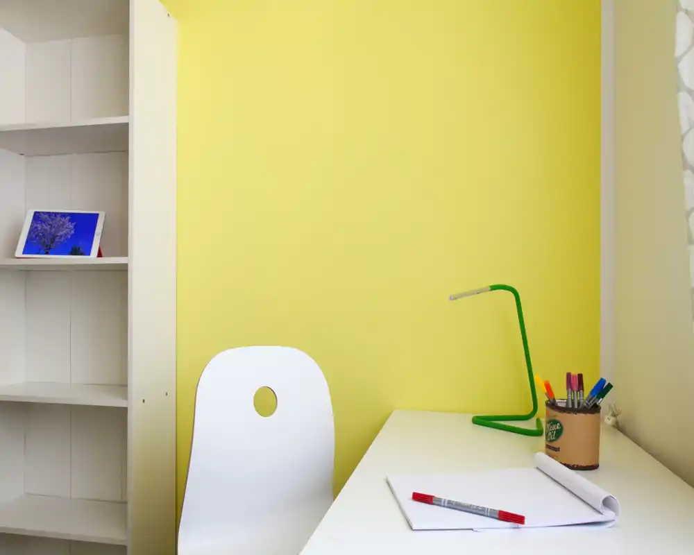 Rincón de trabajo luminoso y minimalista con escritorio y silla blancos frente a una pared amarilla, lámpara verde y material de papelería ordenado sobre la mesa — ideal para una pequeña oficina en casa o zona de estudio.