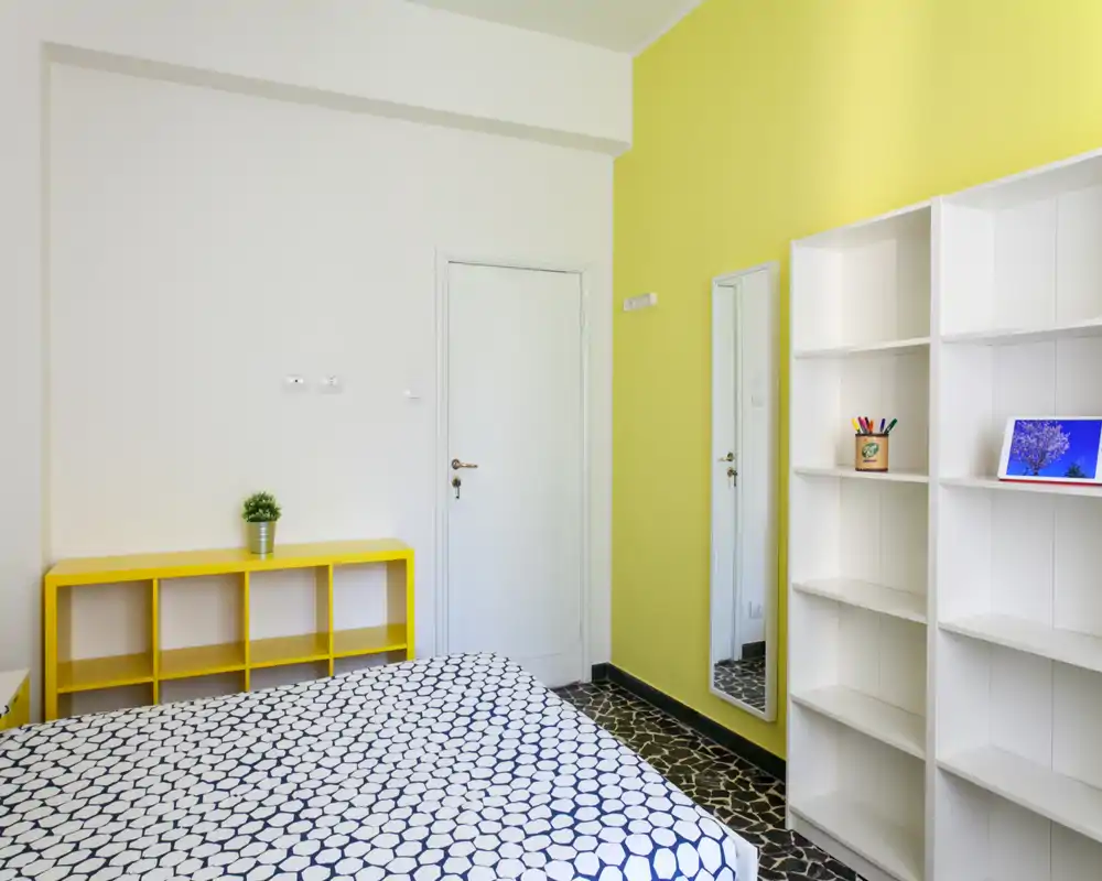 Dormitorio luminoso y minimalista con colcha estampada, estantería amarilla y espejo de cuerpo entero; estilo moderno y limpio, apto para un anuncio de alquiler.