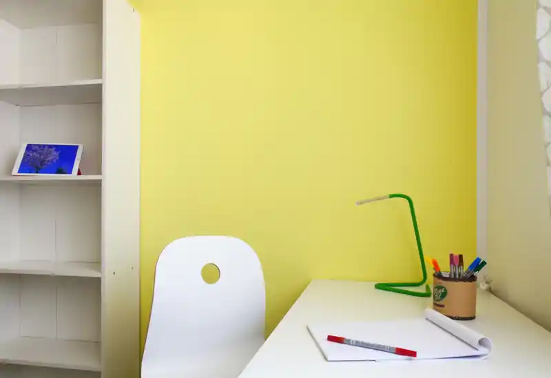 Coin bureau lumineux avec un bureau blanc, une lampe verte et des fournitures sur un mur jaune vif ; chaise blanche et étagères complètent cet espace de travail compact.