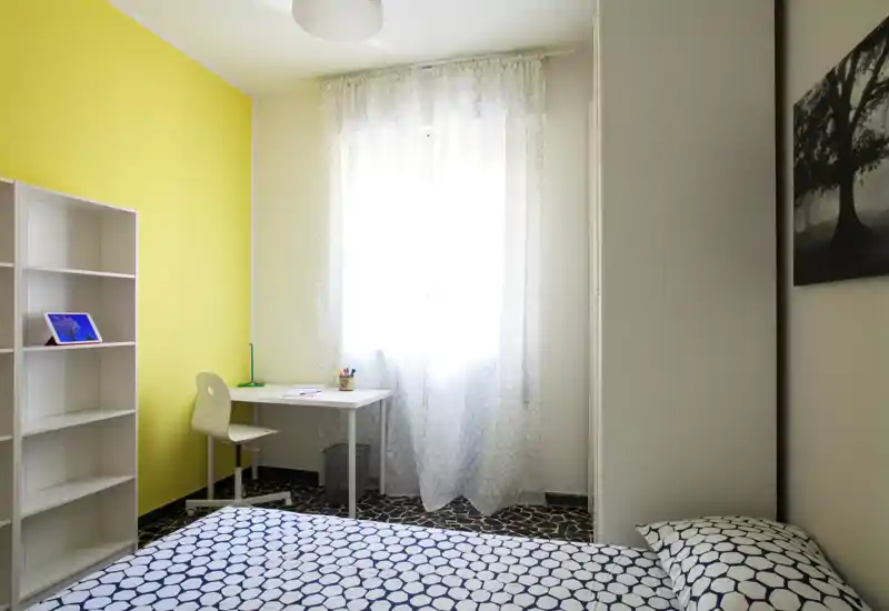 Dormitorio moderno y luminoso con ropa de cama estampada, un pequeño escritorio junto a la ventana y una pared de acento amarilla.