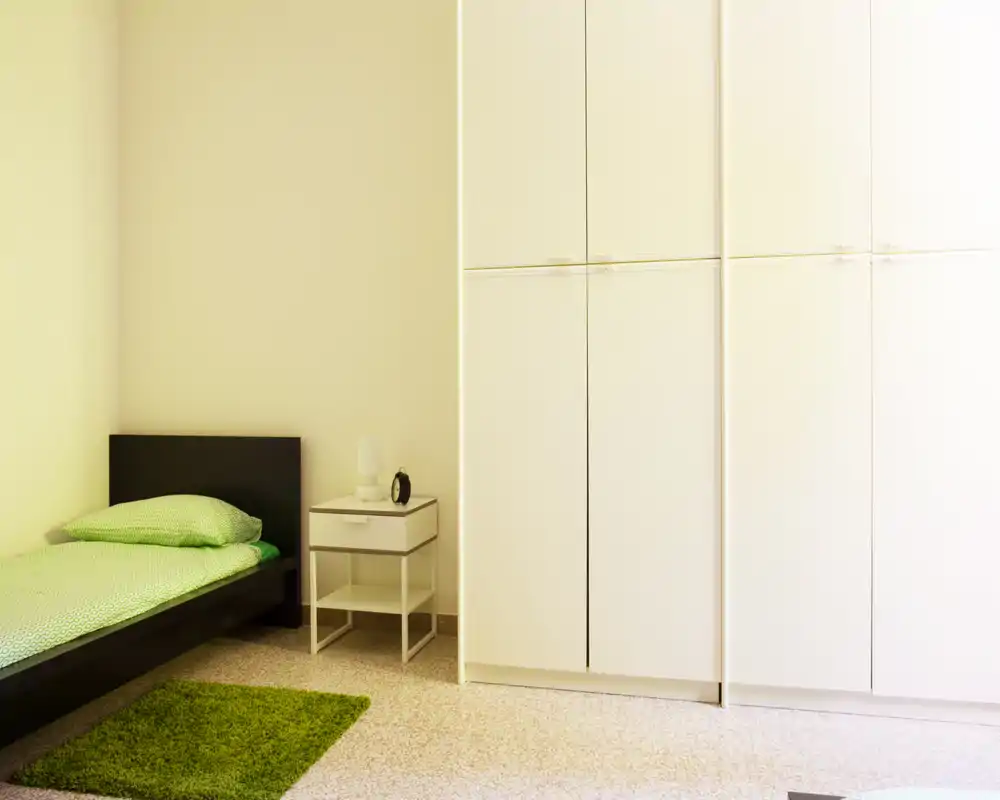 Camera da letto minimal e luminosa con letto singolo nero basso, biancheria verde e armadio a muro bianco; comodino semplice e piccolo tappeto verde per un ambiente ordinato e fresco.