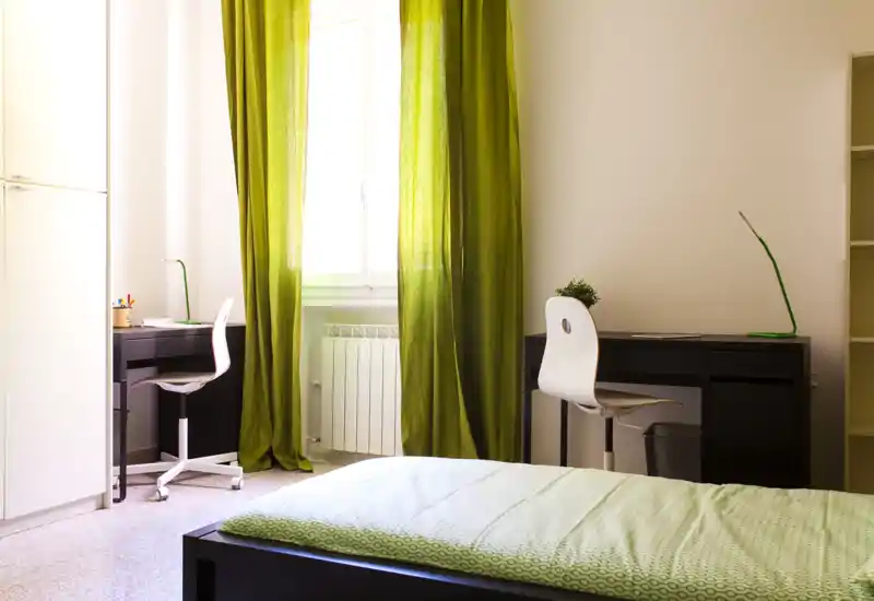 Camera da letto luminosa e moderna con letto ben visibile in primo piano, due scrivanie e tende verdi che lasciano filtrare la luce naturale.