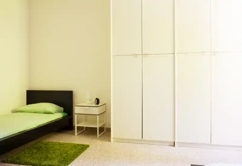 Camera da letto minimale e luminosa con letto singolo, comodino e grande armadio bianco; biancheria verde e piccolo tappeto creano un’atmosfera ordinata e accogliente.