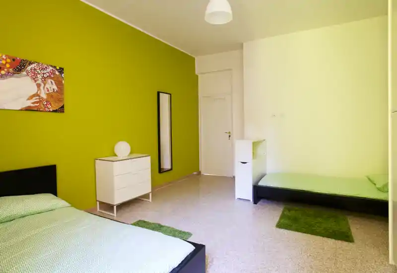 Ampia camera doppia con due letti singoli, parete verde come accento e biancheria coordinata; luminosa, arredata con semplicità, ideale per soggiorni in condivisione.