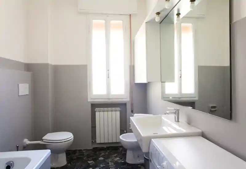 Bright, modern bathroom with a wall-mounted sink, large mirror, toilet, bidet and a bathtub partially visible; neutral grey and white palette with natural light from the window.