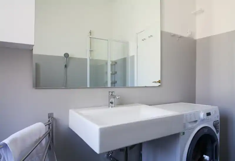 Clean, modern bathroom corner with a large rectangular sink, wall-mounted faucet, mirrored cabinet and a visible shower reflected in the mirror; a washing machine is integrated under the countertop.