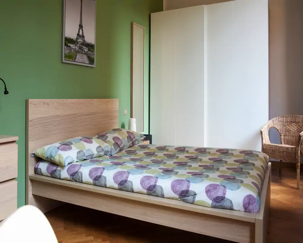 Camera da letto luminosa e ben arredata con letto matrimoniale in legno, copriletto a motivi e un ampio armadio bianco; la luce naturale e il pavimento in legno creano un ambiente accogliente.