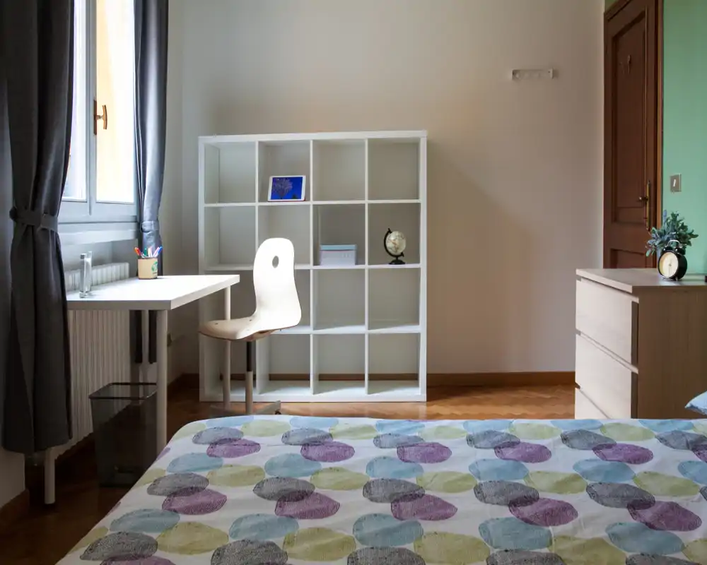 Camera da letto luminosa e ordinata con una coperta a motivi in primo piano, una postazione di lavoro vicino alla finestra e una libreria bianca. La luce naturale e i toni delicati rendono l’ambiente accogliente e funzionale.