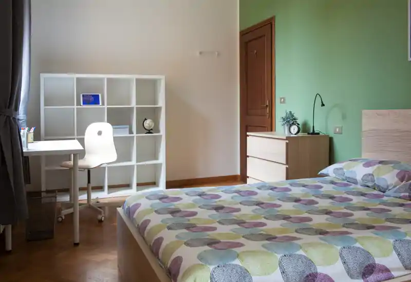Camera luminosa e ben arredata: letto matrimoniale in primo piano con copriletto fantasia, comodino e una scrivania con scaffale vicino alla finestra.
