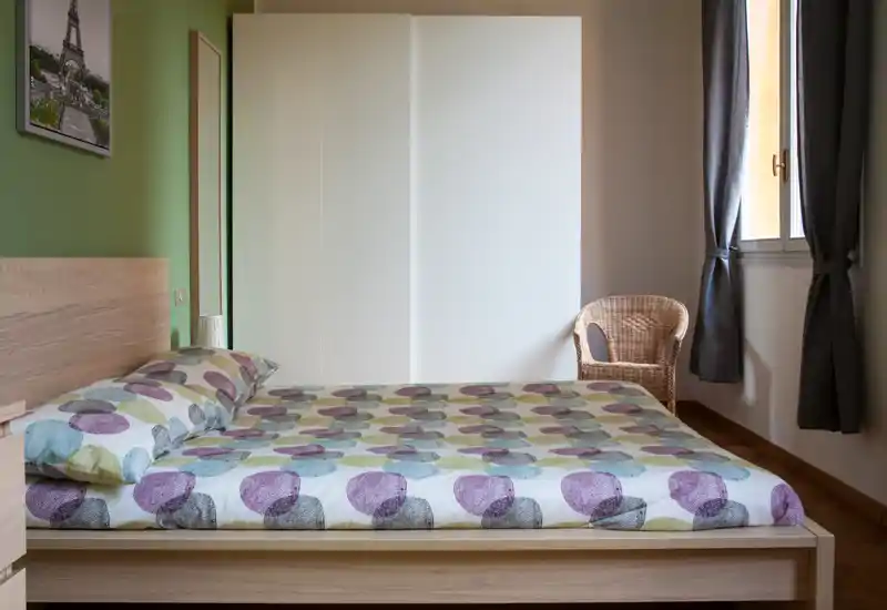 Camera da letto luminosa e ordinata con letto matrimoniale in legno chiaro, un grande armadio bianco e una finestra con tende che lasciano entrare luce naturale.