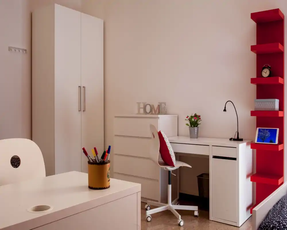 Angolo camera moderno e ordinato con armadio bianco, comò e scrivania, scaffalatura rossa e sedia confortevole — ideale per uno studente o come angolo studio in camera da letto.