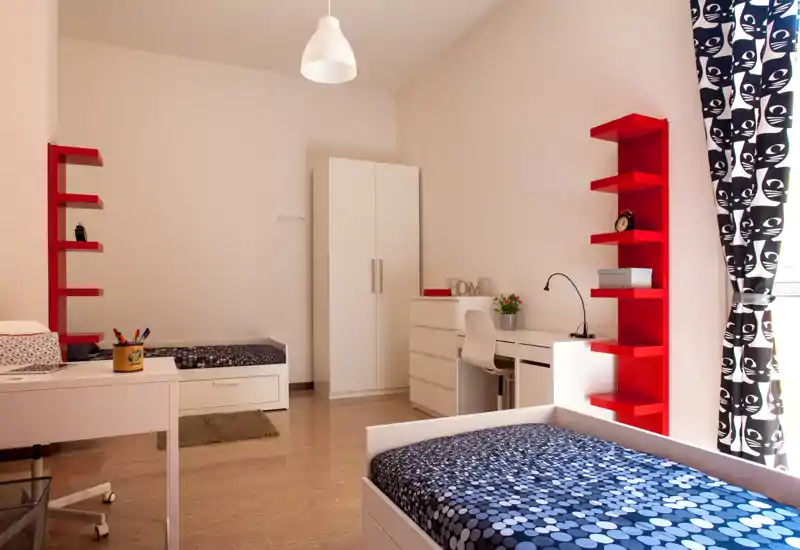 Dormitorio moderno y luminoso con una cama individual estampada en primer plano, una segunda cama y un armario al fondo, además de un escritorio y estanterías rojas para almacenamiento y decoración.