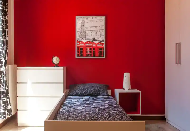 Dormitorio individual compacto con cama bien hecha frente a una pared roja intensa, muebles blancos de almacenamiento y luz natural desde la ventana.