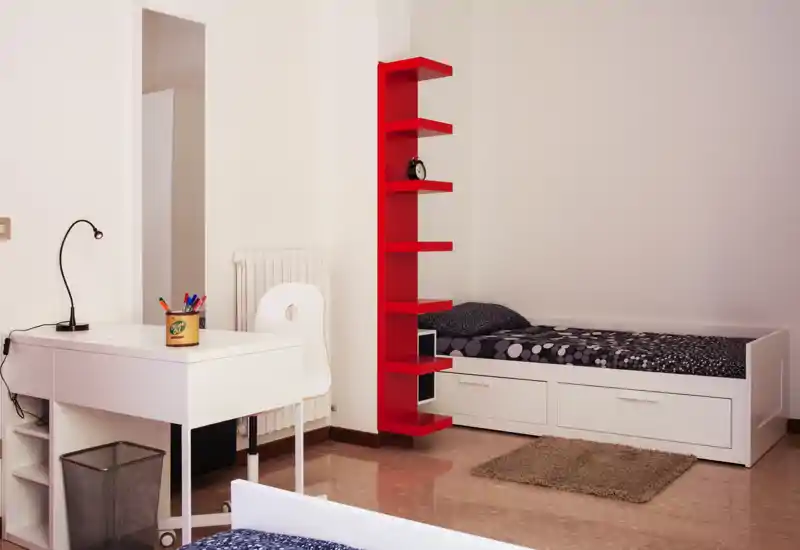 Dormitorio minimalista y luminoso con una cama individual blanca, cajones de almacenamiento, una estantería vertical roja y un pequeño escritorio, ideal para un estudiante o ocupante individual.