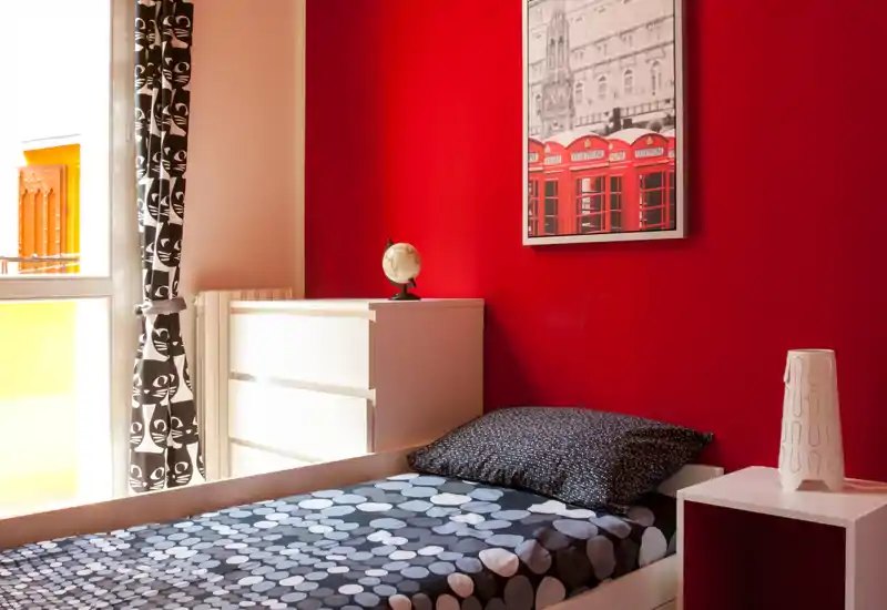 Dormitorio individual luminoso y con estilo, con pared roja de acento, ropa de cama estampada y luz natural de una gran ventana — compacto y acogedor para una persona.