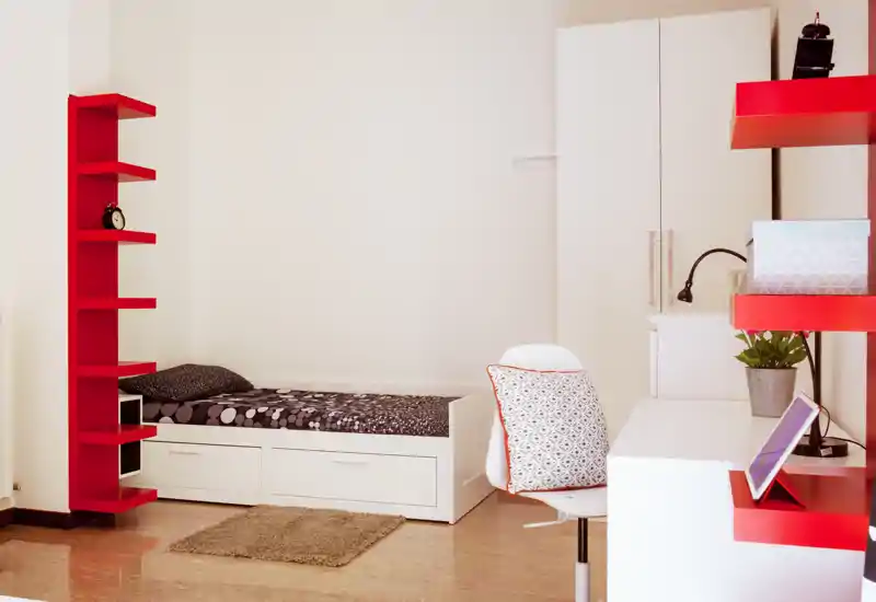 Dormitorio individual moderno y luminoso con almacenamiento bajo la cama, escritorio y estanterías rojas que aportan un acento de estilo.
