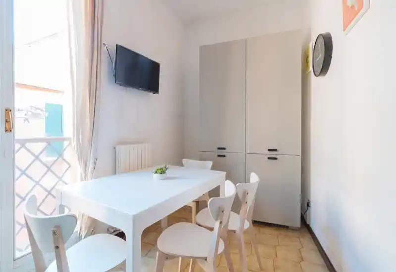 Zona pranzo luminosa e minimal con tavolo bianco e quattro sedie abbinate vicino alla porta del balcone; toni neutri e un armadio compatto creano uno spazio funzionale e arioso.