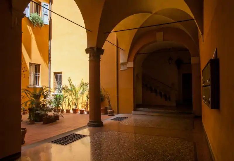 Cortile coperto e soleggiato con pavimento in cotto, vasi di piante e archi a colonnato che portano a una scala; pareti gialle calde che ricordano un’atmosfera mediterranea.