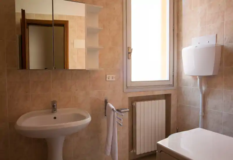 Bagno pulito e ben illuminato con lavabo a colonna, pensile con specchio, porta asciugamani e luce naturale da una finestra — ideale per annunci che valorizzano la funzionalità.