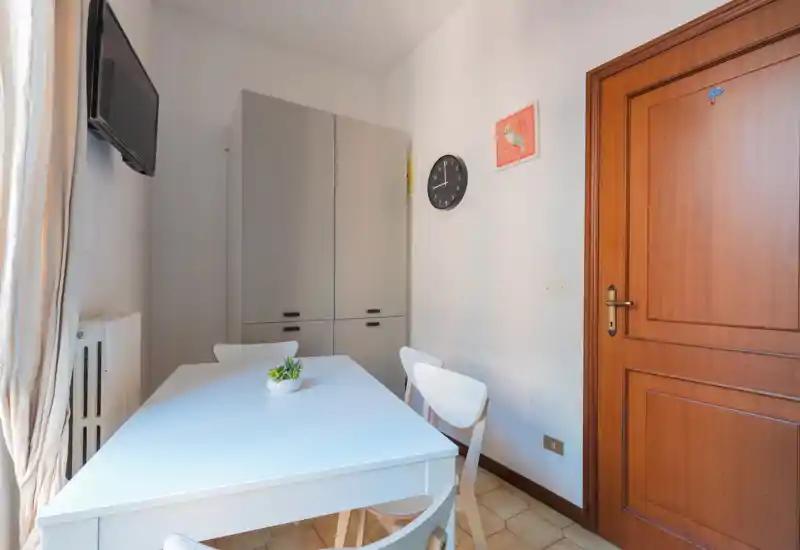 Angolo pranzo compatto con tavolo e sedie bianche, un armadio alto e luce naturale proveniente dalla finestra a sinistra. Arredamento semplice con una piccola pianta al centro e un orologio a parete.