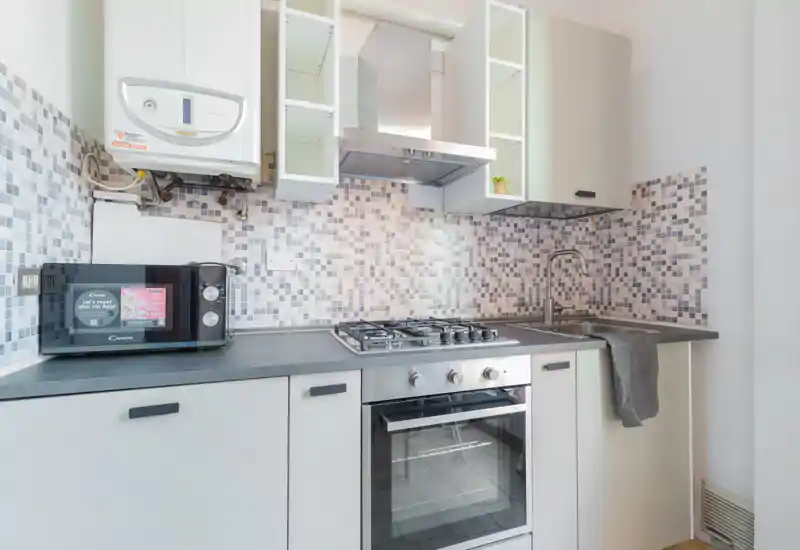 Cucina moderna compatta con piano cottura a gas, forno incorporato, cappa in acciaio e paraschizzi con piastrelle a mosaico; piano con lavello e microonde — ambiente luminoso e ordinato ideale per affitti brevi.