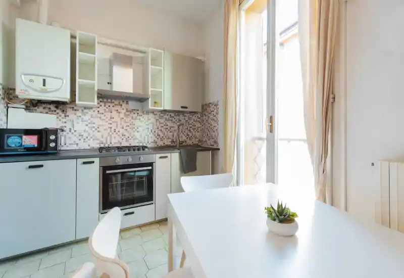 Cucina compatta e luminosa con forno incassato, piano cottura a gas e un piccolo tavolo da pranzo accanto a una grande finestra che illumina l’ambiente.