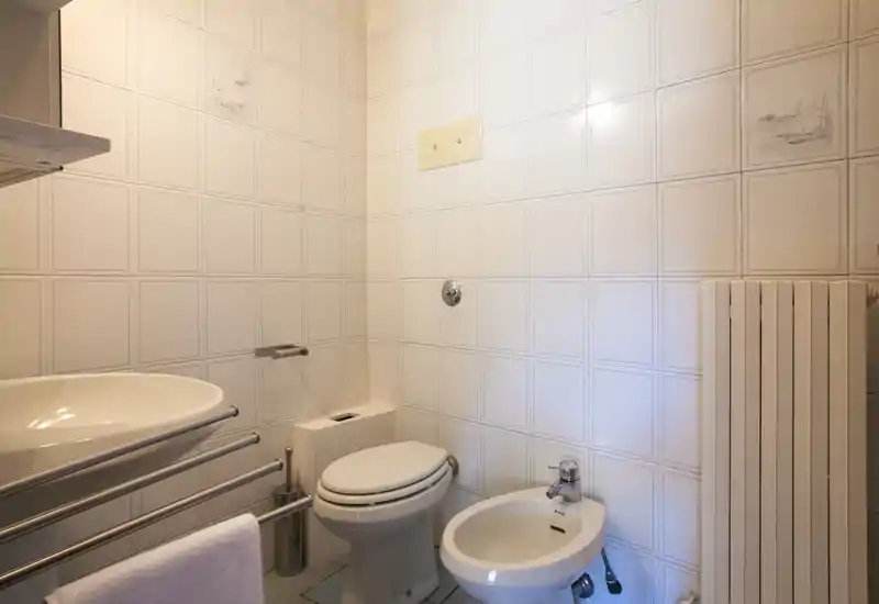 Bagno compatto e luminoso con WC, bidet, lavabo e termosifone; piastrelle bianche e arredi neutri creano uno spazio pulito e funzionale.