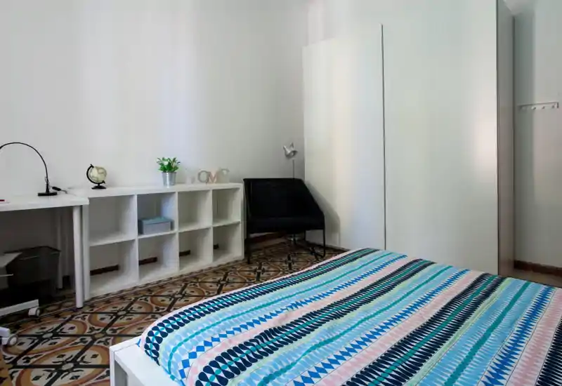 Bright, minimal bedroom with a patterned bedspread, white shelving and a black accent chair — clean, modern and well lit.