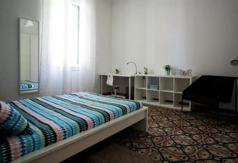 Bright, minimalist bedroom with a low white platform bed dressed in colorful patterned linens, a tall mirror, desk and shelving along the wall, and decorative tiled floor.