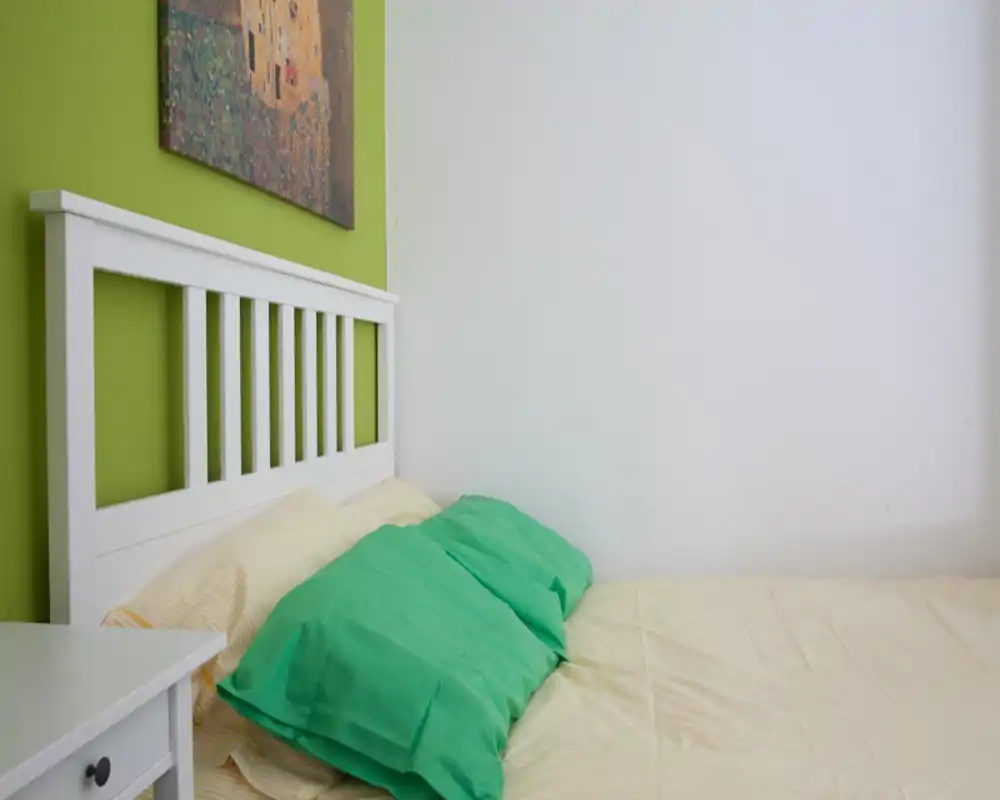 Coin de chambre propre et lumineux avec une tête de lit en bois blanc, des oreillers verts et une literie neutre. La composition met en valeur un lit soigné devant un mur accent vert avec une toile décorative.