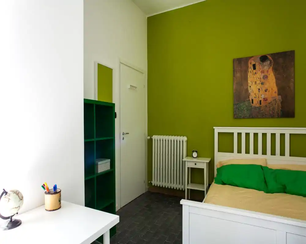Chambre lumineuse et soignée avec un lit en bois blanc, des coussins verts et un mur d'accent vert assorti. Une petite table de chevet, un radiateur et un tableau décoratif complètent l'ambiance moderne et apaisante.