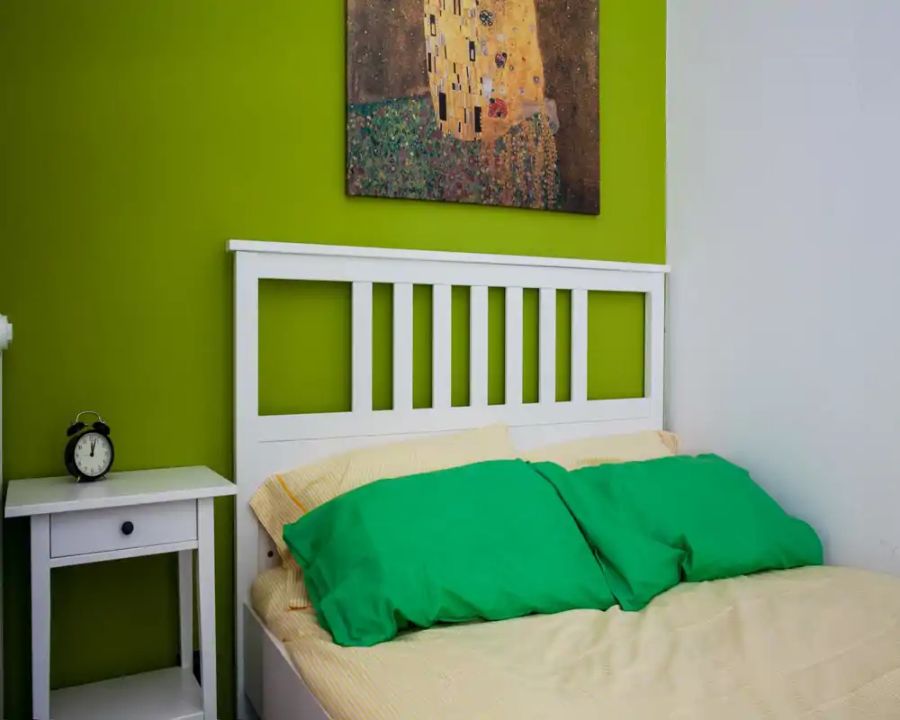 Chambre moderne et lumineuse avec tête de lit blanche à lattes, oreillers verts et table de chevet avec réveil. Le mur d'accent vert vif et le tableau apportent une atmosphère fraîche et accueillante.