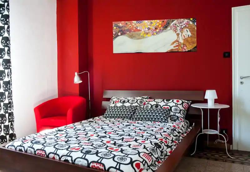 Bright, stylish bedroom with a double bed dressed in bold patterned linens, a red accent wall and a cozy red armchair by the window.