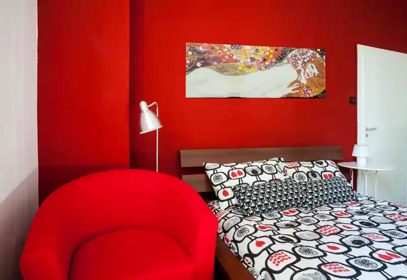 Bright, stylish bedroom with bold red walls and patterned bedding; wooden headboard, bedside table and a comfortable red armchair create a modern, cozy atmosphere.
