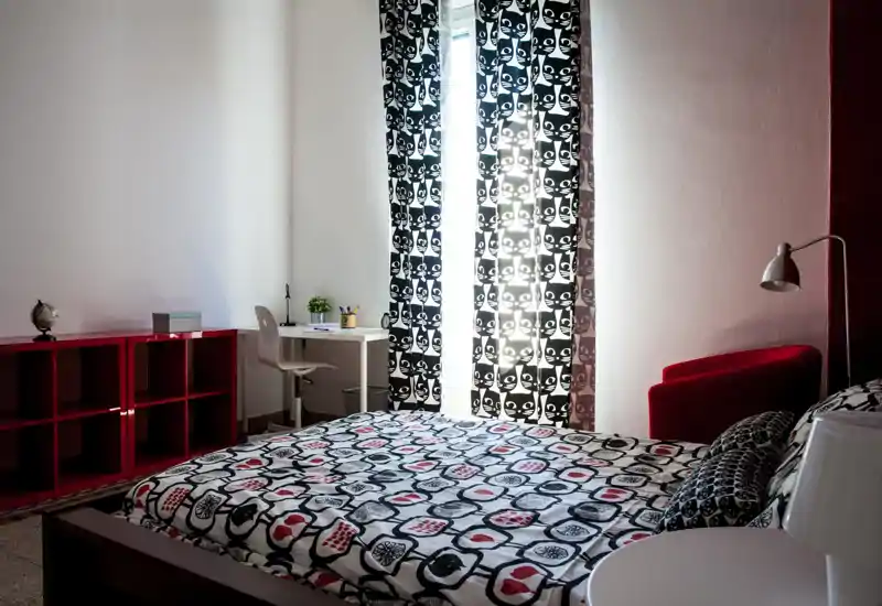 Bright, stylish bedroom with a patterned double bed in the foreground, a small desk by the window and a red armchair. The room features bold black-and-white curtains and modern, playful textiles.