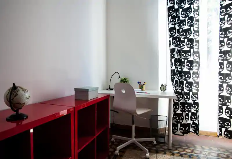 Bright and tidy living area corner used as a home office: a white desk with a chair, small lamp, plant and stationery beside large patterned curtains and a glossy red shelving unit.