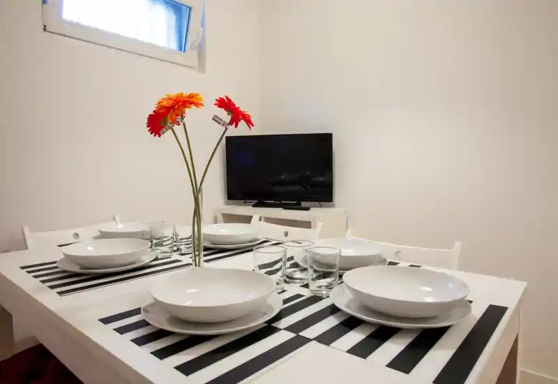 Bright, minimalist dining area with a table set for four, striped placemats and a simple floral centerpiece; clean white furnishings and a small TV in the background.