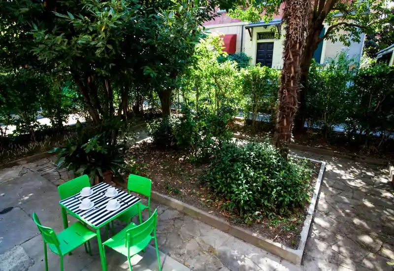 Sunny private garden/terrace with a small table and four bright green chairs set on a stone patio, surrounded by leafy hedges and trees — ideal for outdoor dining and relaxation.