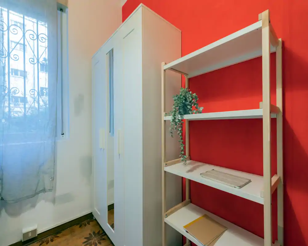 Small bedroom corner with a white wardrobe and open shelving against a warm red accent wall; natural light filters through a nearby window creating a bright, tidy feel.