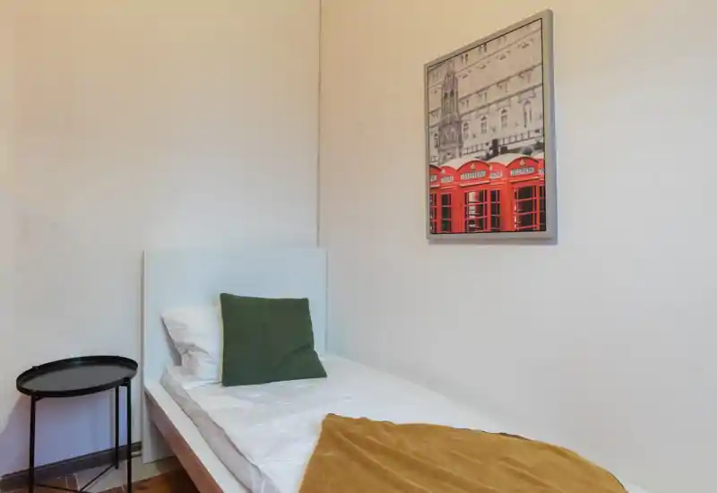 Compact single bedroom with a clean white bed, decorative green cushion and warm throw; simple bedside table and framed artwork create a tidy, welcoming space.