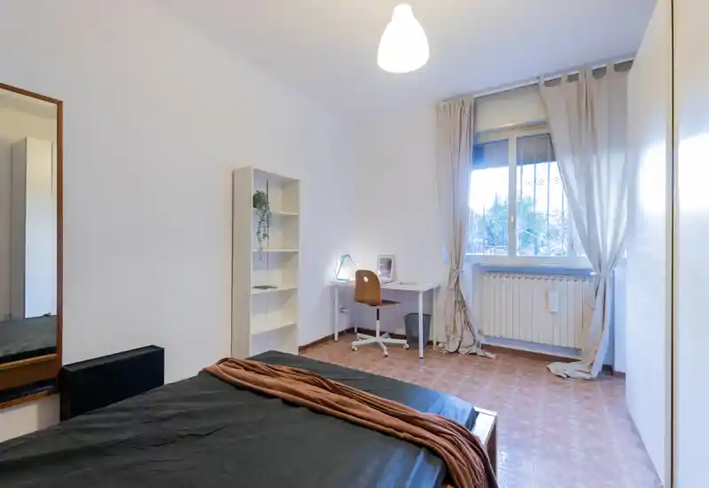 Camera da letto luminosa e minimale con letto matrimoniale in primo piano, scrivania vicino alla finestra, libreria e arredi neutri che creano un ambiente funzionale e tranquillo.