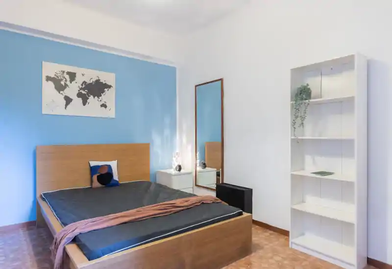 Camera da letto luminosa e dal design semplice con letto matrimoniale in legno contro una parete blu, comodino e specchio a figura intera. Linee pulite e scaffalatura minimal creano un ambiente moderno e rilassante.