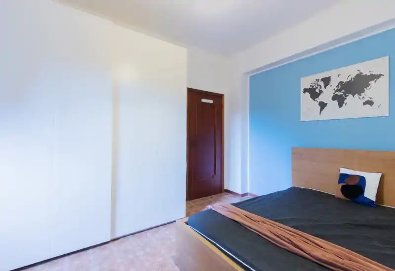 Camera da letto luminosa e minimal con testiera in legno su parete azzurra, ampia armadiatura bianca e una mappa decorativa: linee pulite e disposizione funzionale.