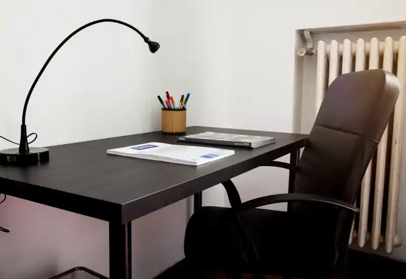 Pequeño espacio de trabajo ordenado con escritorio oscuro, lámpara de sobremesa flexible, portalápices y una cómoda silla de oficina de cuero junto al radiador — ideal para concentrarse en el trabajo.