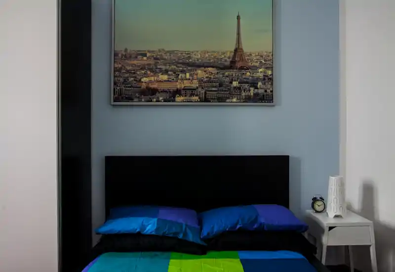 Dormitorio moderno y luminoso con cama doble vestida con ropa de cama colorida, cabecero sencillo y un cuadro de la ciudad colgado encima. Una mesita con lámpara y reloj completan la escena ordenada y acogedora.