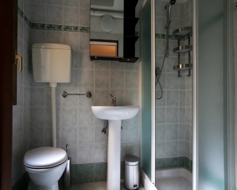 Bagno compatto e ben illuminato con lavabo a colonna, WC con cassetta a muro e box doccia angolare con porte in vetro satinato. Piastrelle neutre e rubinetteria cromata donano un ambiente pulito e funzionale, perfetto per affitti brevi.