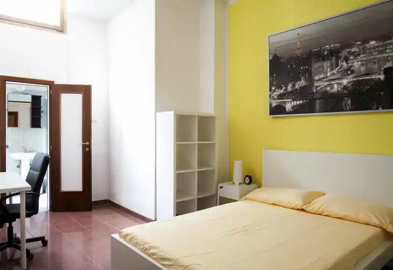 Camera da letto luminosa e ordinata con letto matrimoniale, comodino e scaffalatura; parete gialla e grande foto incorniciata donano vivacità.